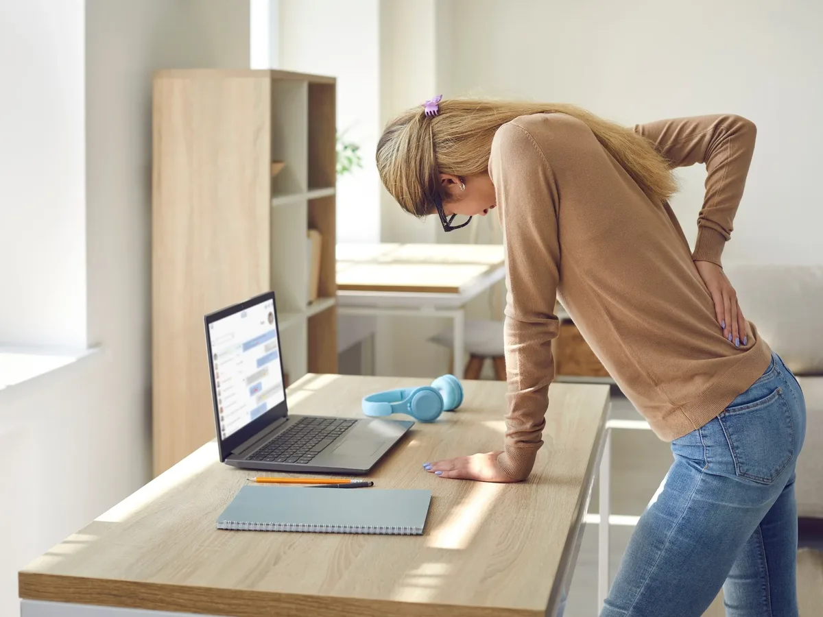Woman leaning on her desk with one hand pressed to her lower back